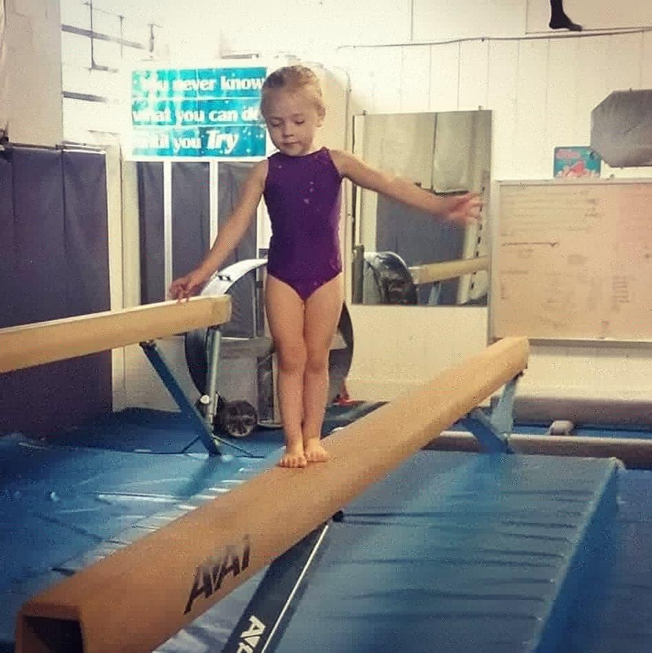young girl with blonde hair in a purple outfit balancing on a beam